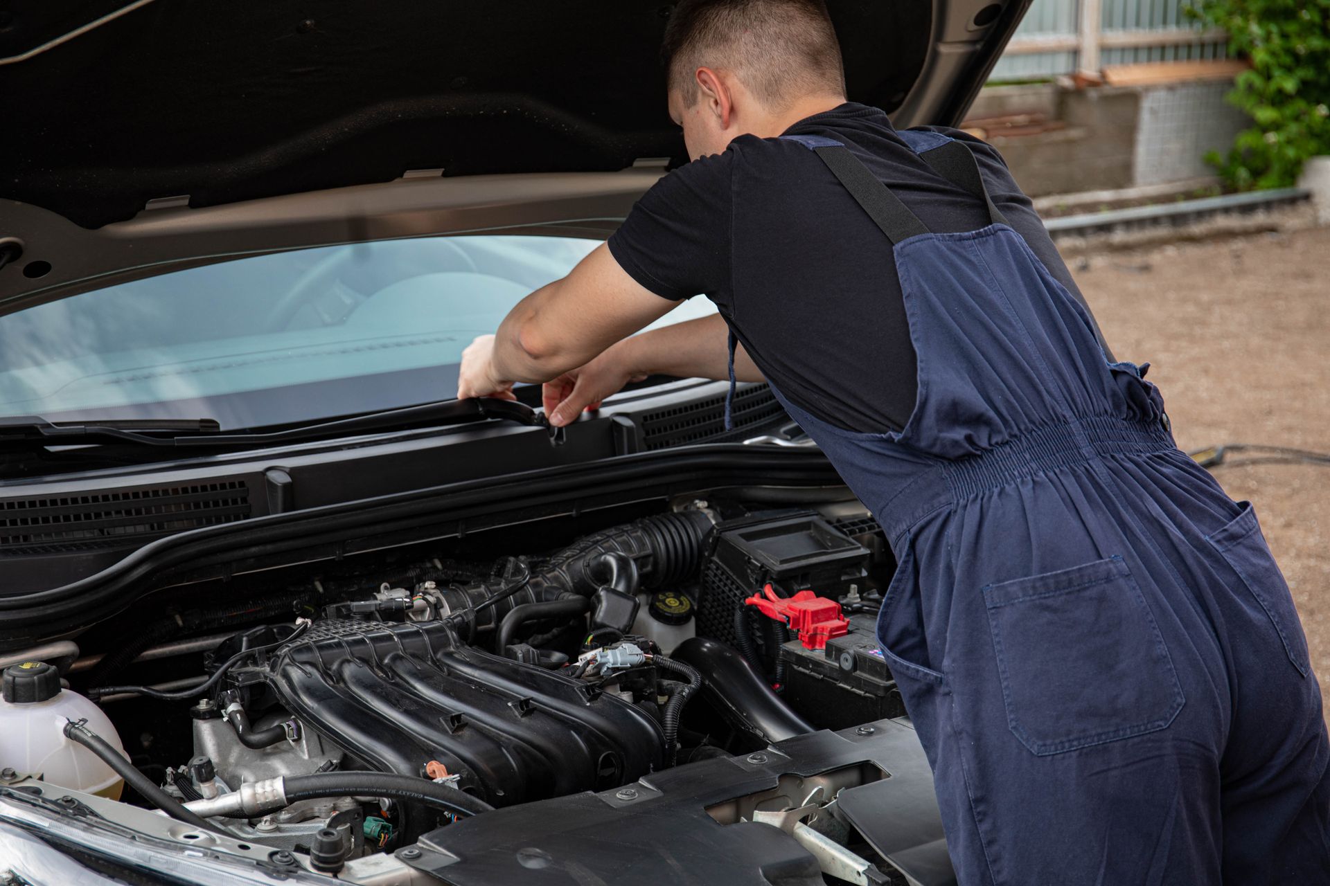 A mechanic leaning into an open hood, working outdoors on gravel rather than in a shop bay